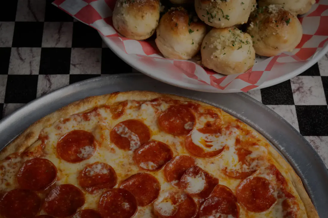 A pepperoni pizza and a basket of garlic knots on the checkered cloth